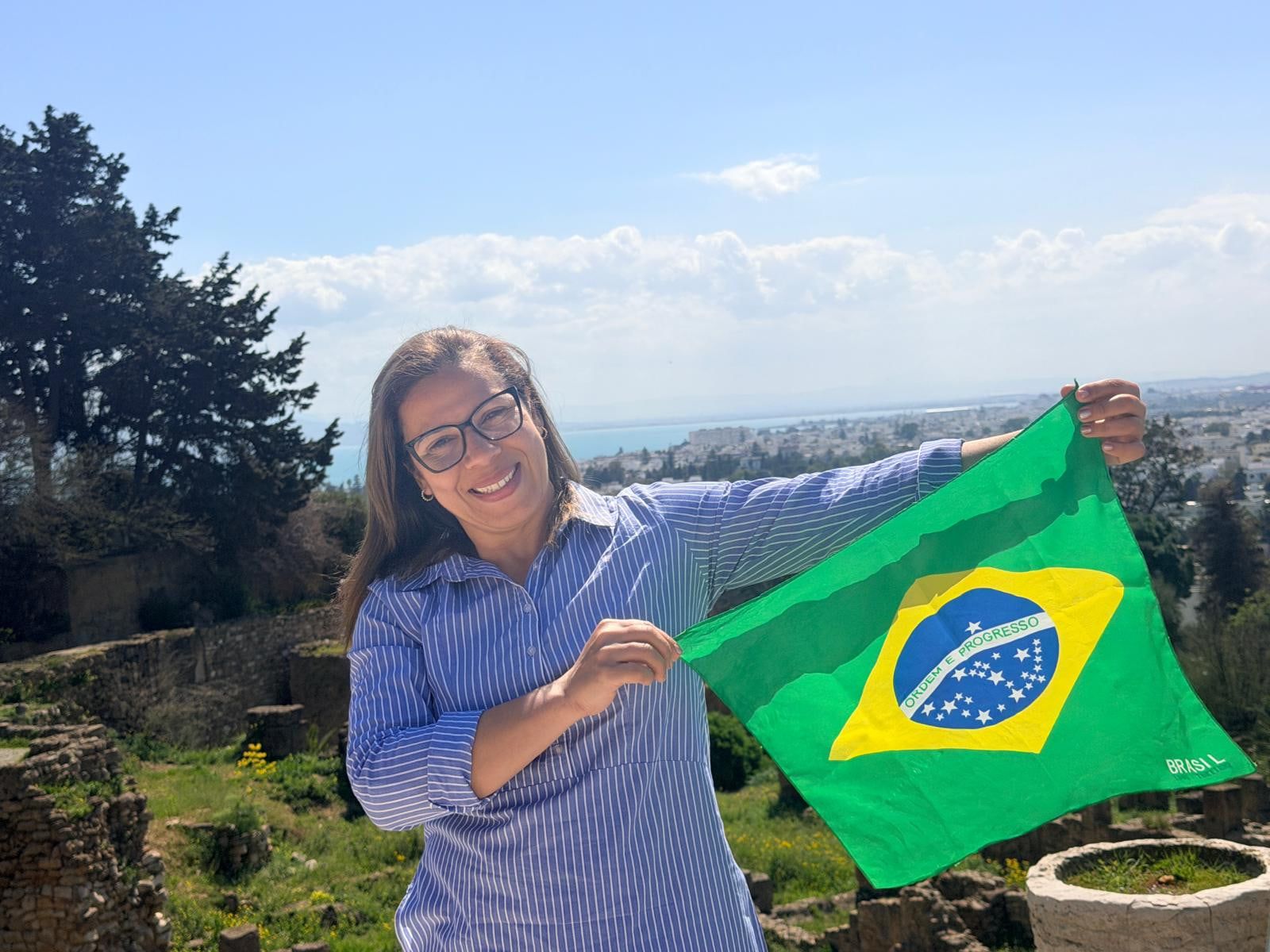Érika sorrindo com a bandeira do Brasil nas ruínas de Cartago, com a cidade e o mar ao fundo